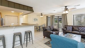 A modern living room with a brown leather sofa, a blue armchair, and a coffee table. There is a kitchen area with a bar counter and stools. The room has a ceiling fan and a large window with curtains.