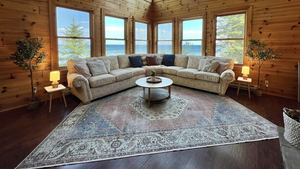 A cozy cabin interior with a large sectional sofa, a round coffee table, and a patterned rug. There are multiple windows providing a view of the outdoors. - Private & secluded beach front on Lake Huron! (Cheboygan)