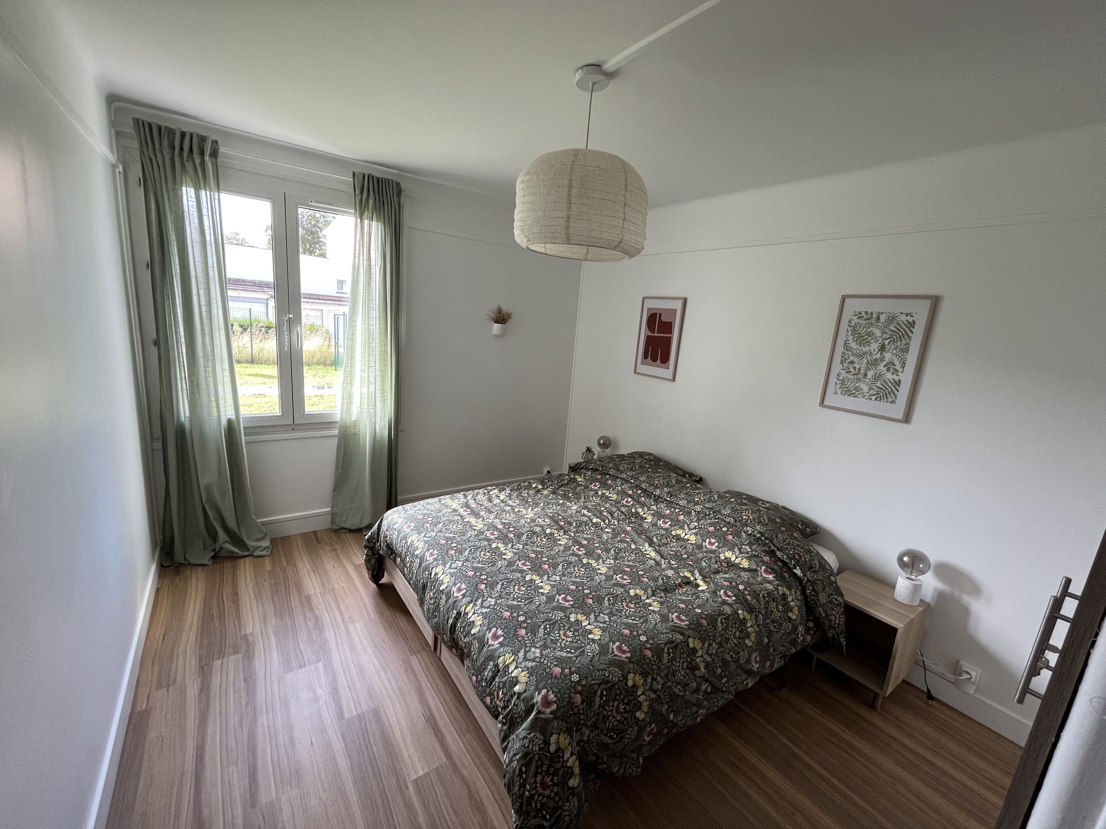 A bedroom with a bed, a bedside table, a hanging light fixture, and a window with curtains.