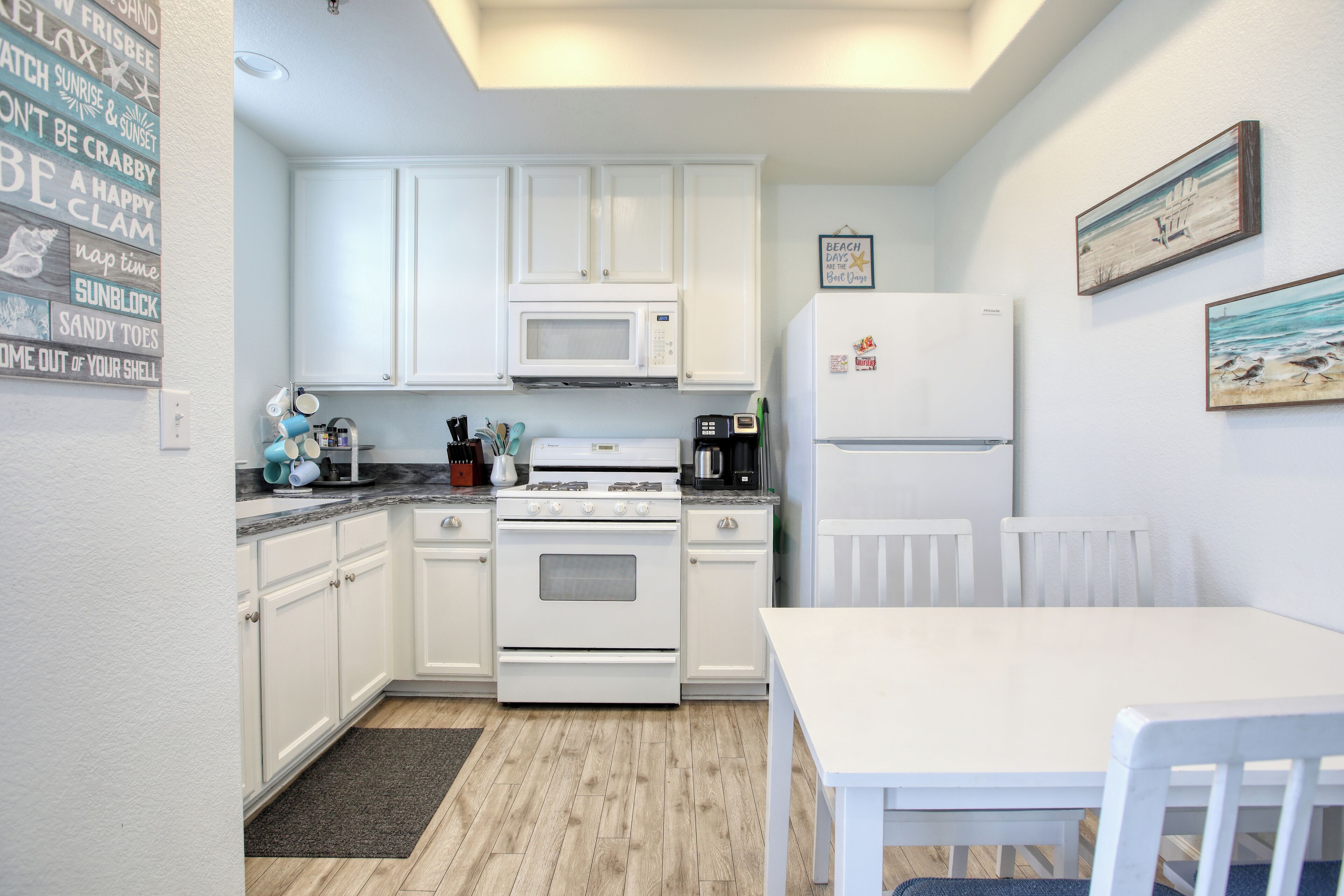 Une cuisine compacte avec des meubles blancs, un four à micro-ondes, une cuisinière et un réfrigérateur. On y trouve une table à manger et des chaises, ainsi que des œuvres d’art accrochées aux murs.