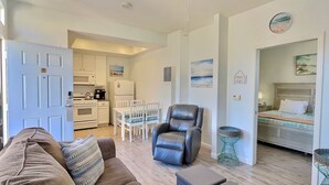 Un espacio habitable compacto con kitchenette, zona de comedor, sala de estar con sofá y reposapiés, y dormitorio con cama y mesita de noche.