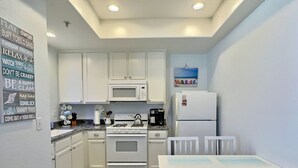 Cocina compacta con gabinetes blancos, microondas, cocina a gas y refrigerador. Cuenta con una pequeña mesa de comedor con sillas.