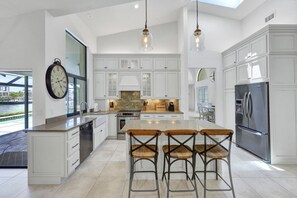 A spacious kitchen with white cabinetry, a large island with bar stools, a stainless steel refrigerator, and a clock on the wall.