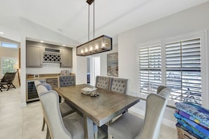 Un coin repas avec une table en bois et des chaises rembourrées, une cuisine équipée d’appareils modernes et une grande fenêtre avec des stores.