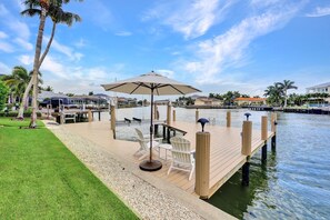 A wooden dock extending into calm water, with a grassy area and palm trees on the shore.