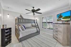 A bedroom with a bunk bed, a ceiling fan, a TV, a dresser, and a window with blinds.