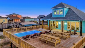 Une terrasse autour de la piscine, avec des chaises longues et des tables de pique-nique, un espace de terrasse couvert et une maison à l’extérieur bleue et au toit en pente.