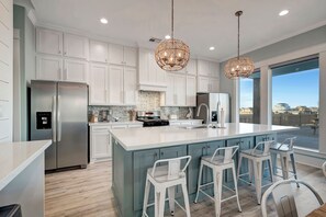 A modern kitchen with white cabinets, a large island with bar stools, stainless steel appliances, and a view through large windows.