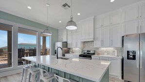 A modern kitchen with white cabinets, a large island, stainless steel appliances, and a view through large windows. - Sunny & 75: 5  BR, 5.5  BA House in Crystal Beach, Sleeps 26 (Bolivar Peninsula)