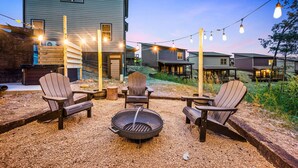 Eine Terrasse mit Adirondack-Stühlen, einem Feuerkorb und Lichterketten vor einer Häuserkulisse.