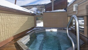 A hot tub with a wooden deck and metal railings, surrounded by snow-covered buildings and a mountainous landscape. - Newly remodeled condo just steps from the gondola, hot tub, sleeps 4! (Vail)