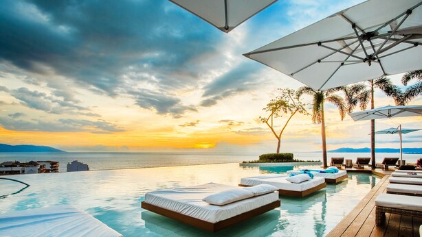 Une piscine à débordement avec des chaises longues et des parasols, offrant une vue imprenable sur le coucher de soleil sur la mer.
