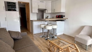 Una sala de estar moderna con un sofá marrón, una mesa de centro de madera y un sillón gris. Se ve una cocina al fondo con armarios blancos y un electrodoméstico rojo.