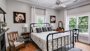 A bedroom with a bed, a chair, a nightstand, a lamp, and two windows.