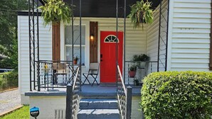 A two-story house with a red front door, a covered porch with outdoor furniture, and a small staircase leading to the entrance. - Cozy home, close to dining, entertaining, military bases, beaches and more. (North Charleston)