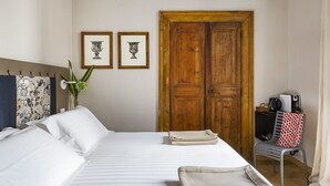 Ett hotellrum med en säng, en trädörr, två inramade tavlor, en stol och ett litet bord med en kaffebryggare.