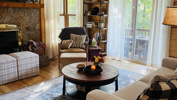 A cozy living room with a fireplace, wooden walls, a ceiling fan, and a large window with curtains.
