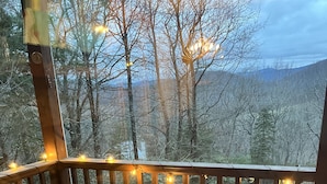 A wooden deck with a table and benches, string lights, and a view of a forested area.