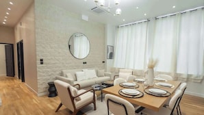 A modern dining area with a wooden table set for four, surrounded by white chairs. A round mirror hangs on the wall, and the room features a light-colored stone wall and wooden flooring. - Spacious Air-conditioned Apartment Center of Paris (Paris)