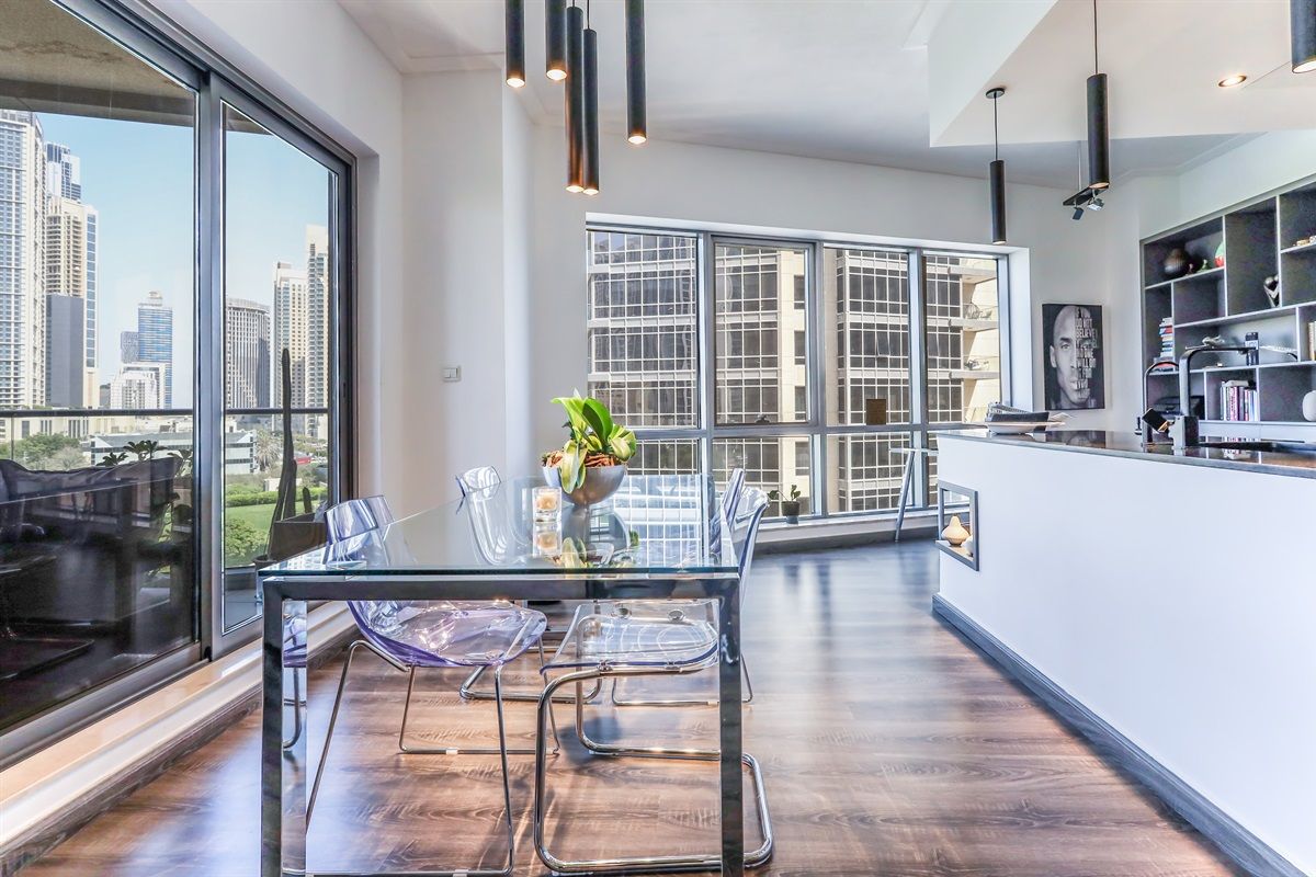 A modern dining area with a glass table and chairs, a city view through large windows, and a kitchen area with a black countertop and shelves.