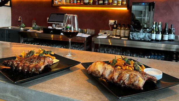 Zona de bar con diversas botellas de licor y vasos en estantes, una copa de vino con vino tinto, y platos de pato asado con vegetales.