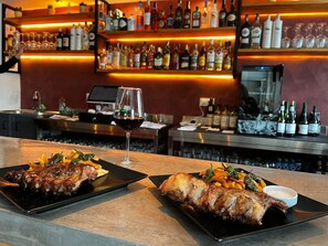 Angolo bar con diverse bottiglie di liquori e bicchieri sugli scaffali, un bicchiere da vino con vino rosso e piatti di anatra arrosto con verdure.