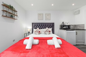 A modern hotel room with a red bedspread, a tufted headboard, and a small kitchenette.