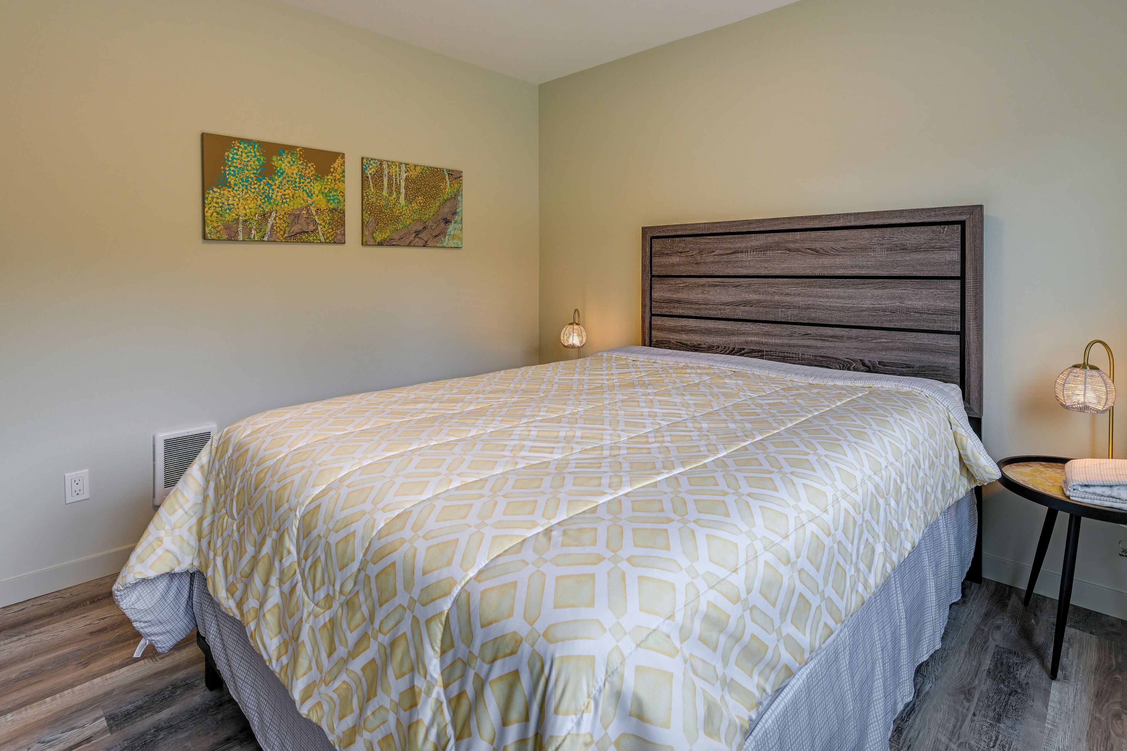 A bedroom with a large bed, a nightstand with a lamp, and two wall-mounted paintings.