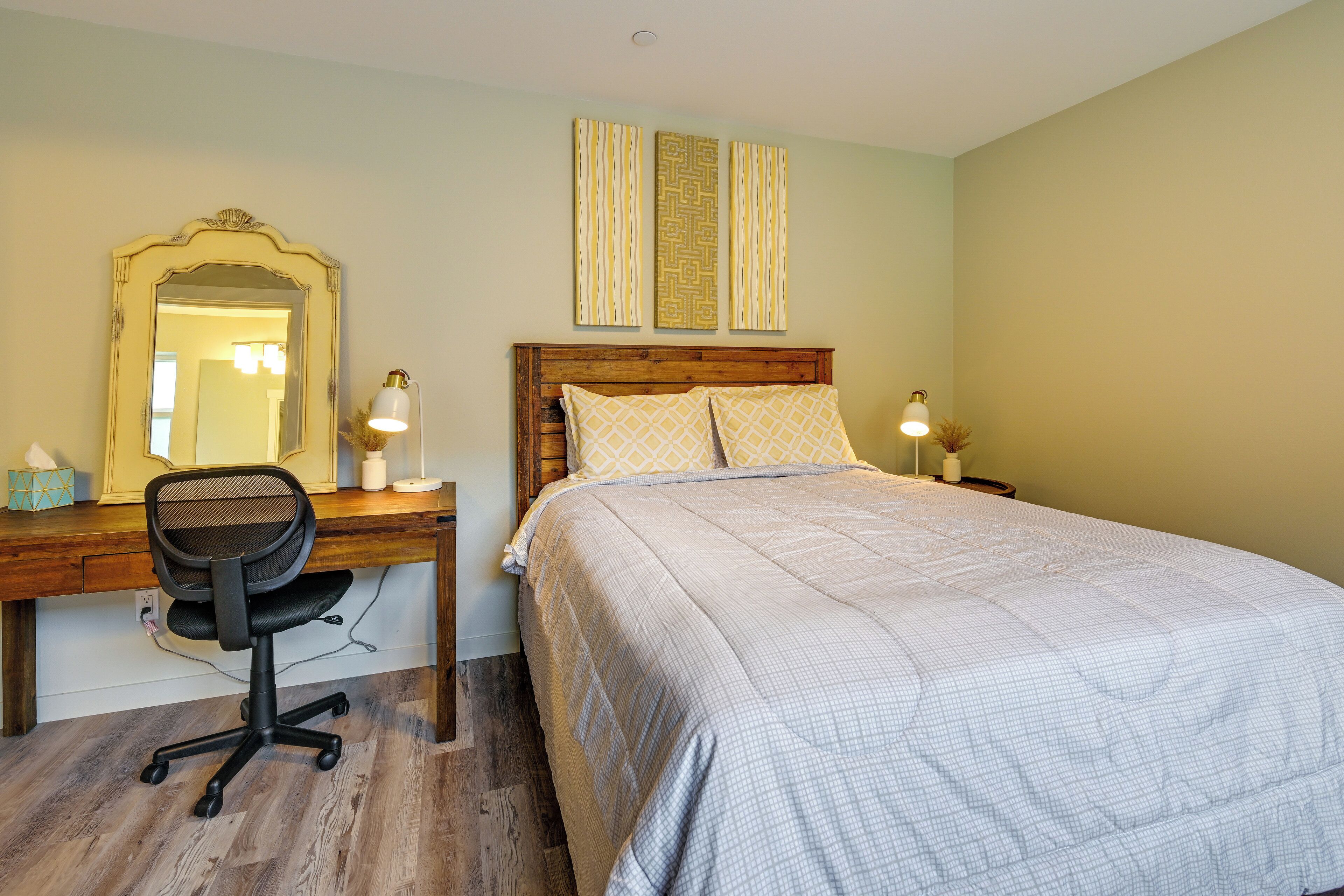 Une chambre avec un bureau en bois, une chaise, un lit recouvert d’une couvre-lit à motifs, une table de chevet avec une lampe et un miroir accroché au mur.