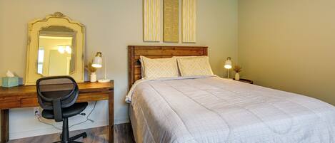 Une chambre avec un bureau en bois, une chaise, un lit recouvert d’une couvre-lit à motifs, une table de chevet avec une lampe et un miroir accroché au mur.