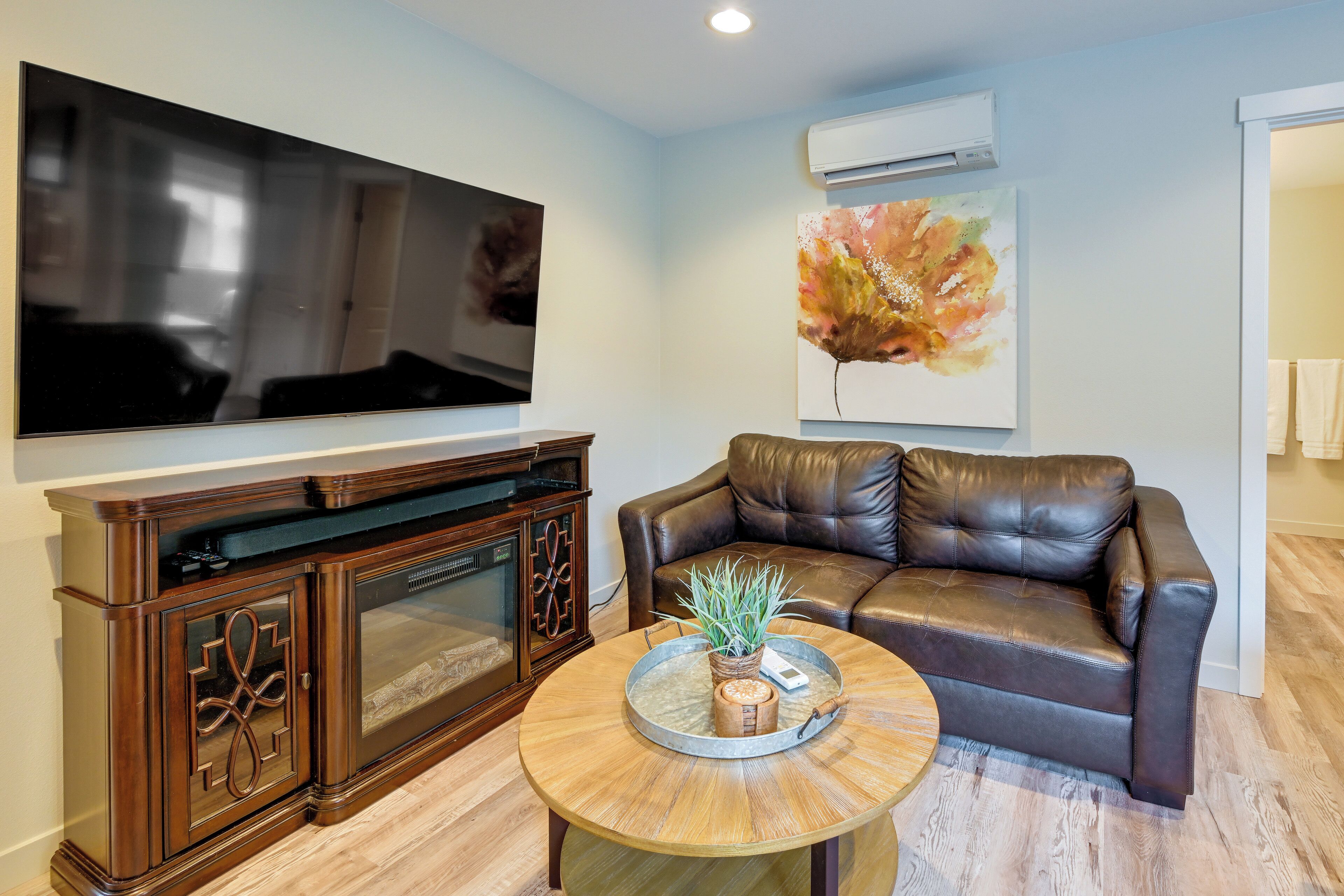 A living room with a flat-screen TV, a brown leather sofa, a coffee table with a plant, and a painting on the wall.