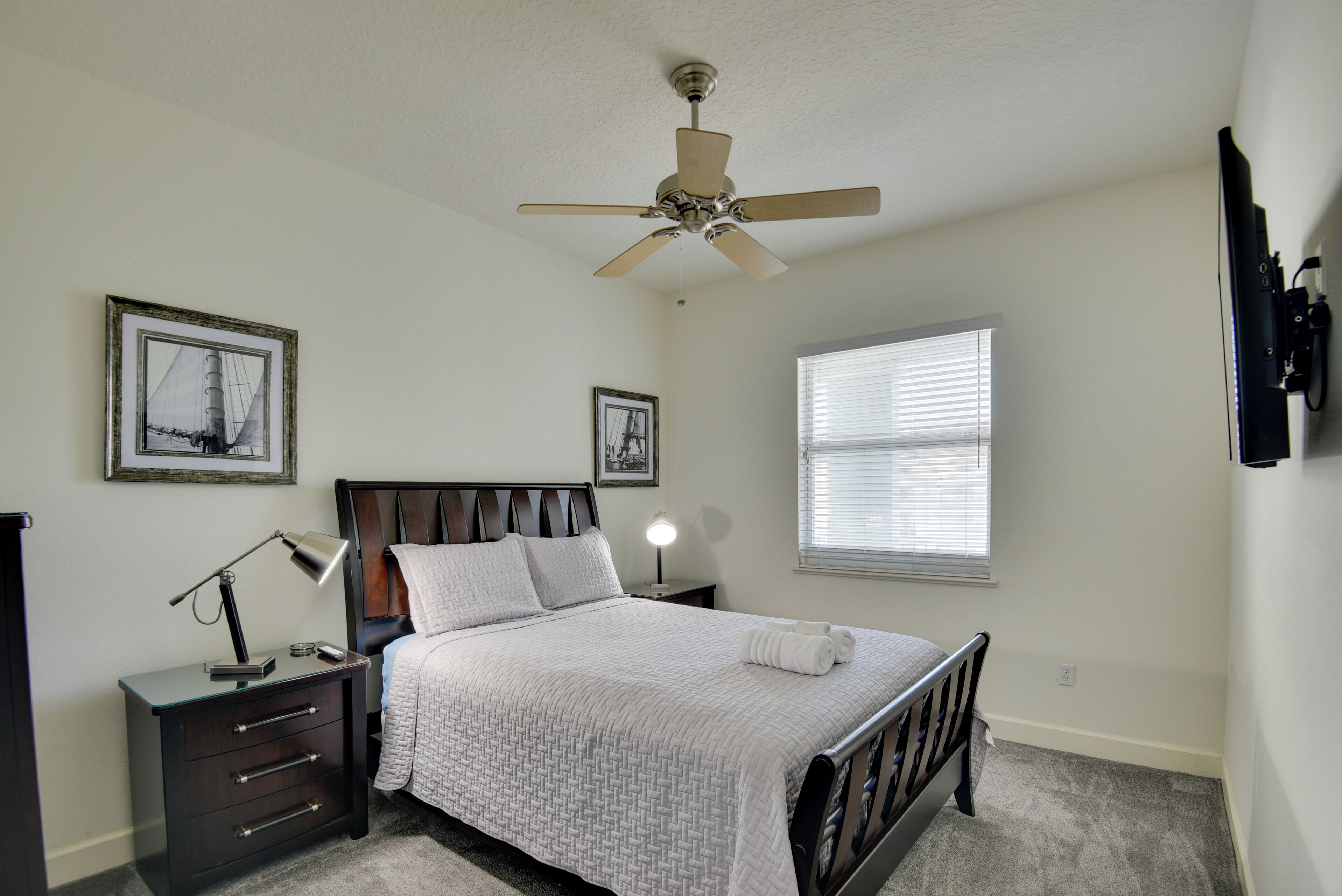 A bedroom with a bed, a nightstand, a desk lamp, a ceiling fan, and two framed pictures on the wall.