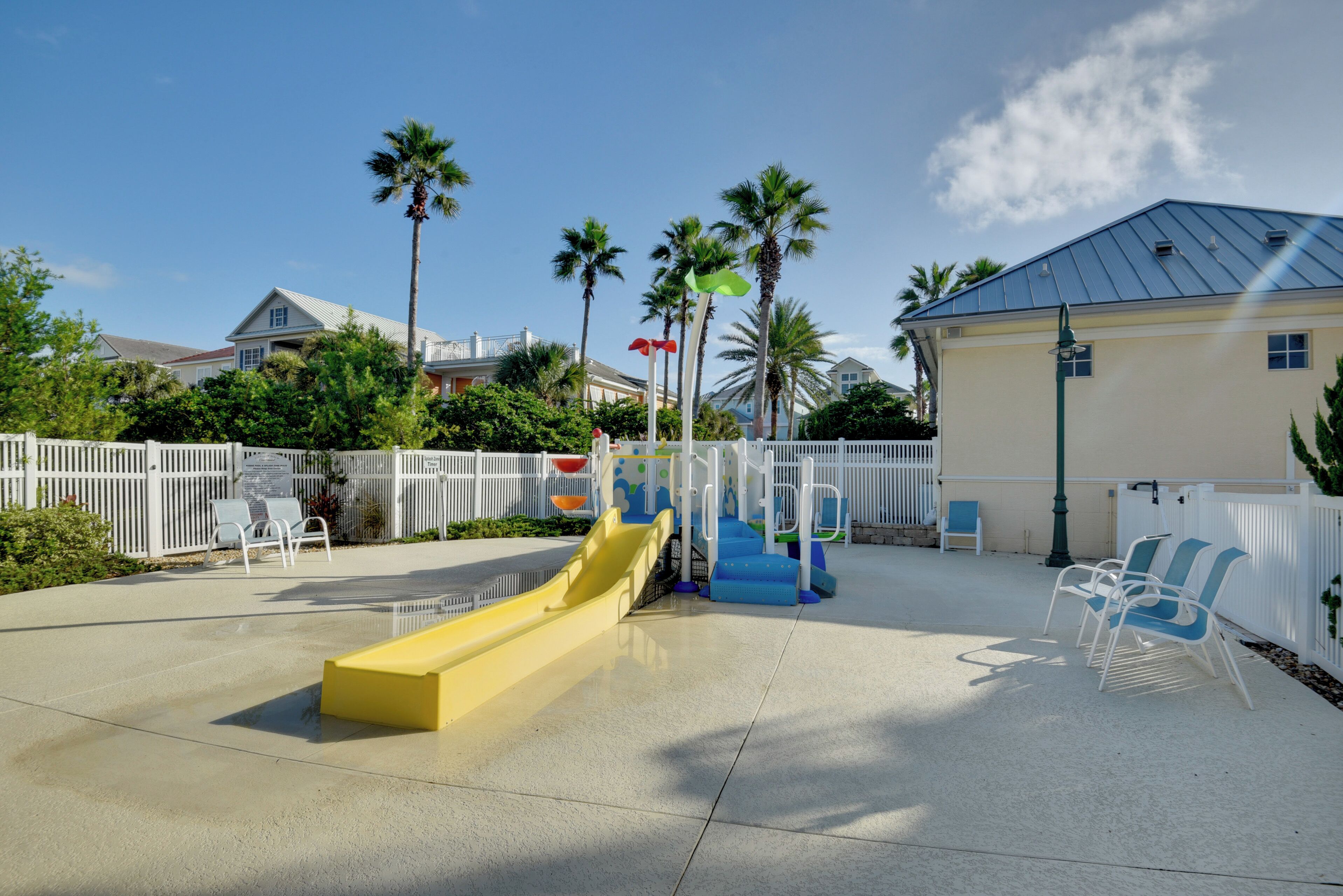 A playground with a yellow slide, blue play structures, and white chairs.