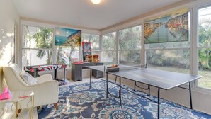 A room with a ping pong table, a white armchair, a desk with a computer, and large windows with tropical views.