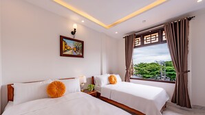 A hotel room with two beds, a painting on the wall, a window with curtains, and a view of greenery outside. - Golden Sun Hotel Hoi An (Da Nang)