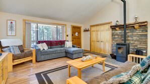 A cozy living room with a stone fireplace, a wooden ceiling fan, and a large window with a view of trees.