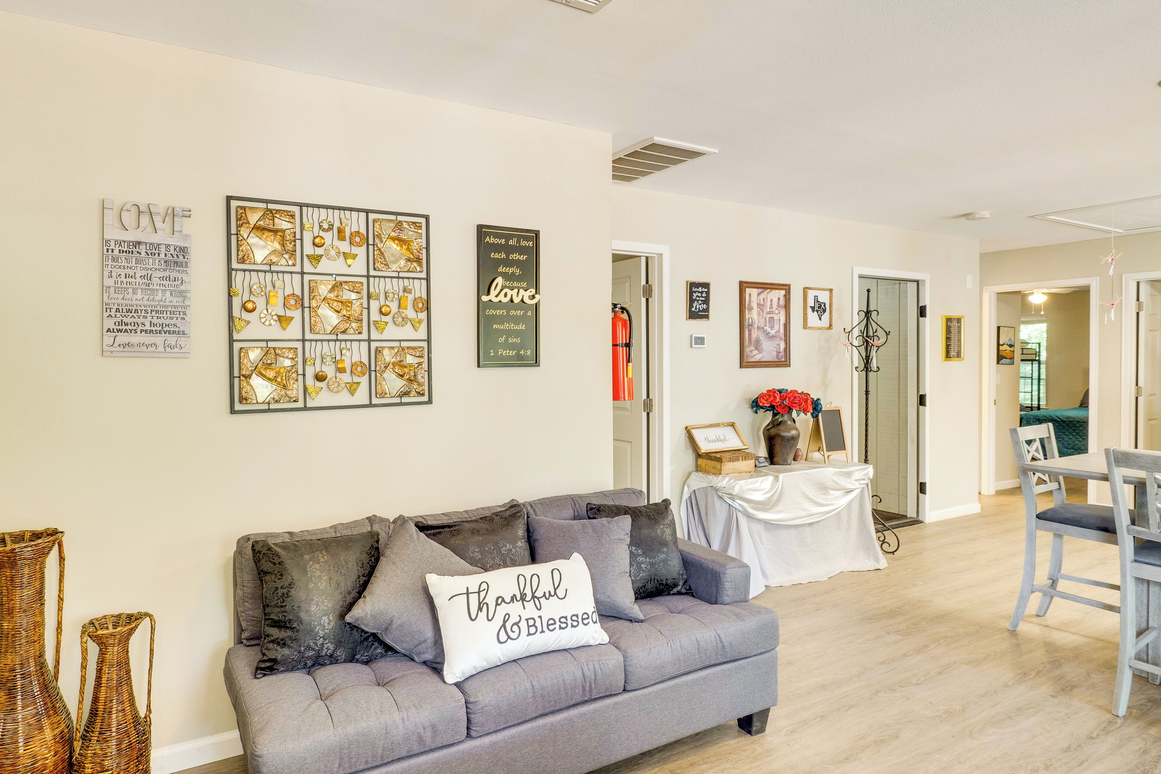 Een woonkamer met een grijze bank, een tafel met een witte tafelkleed, en diverse wanddecoraties, waaronder lijstjes met citaten en kunstwerken.