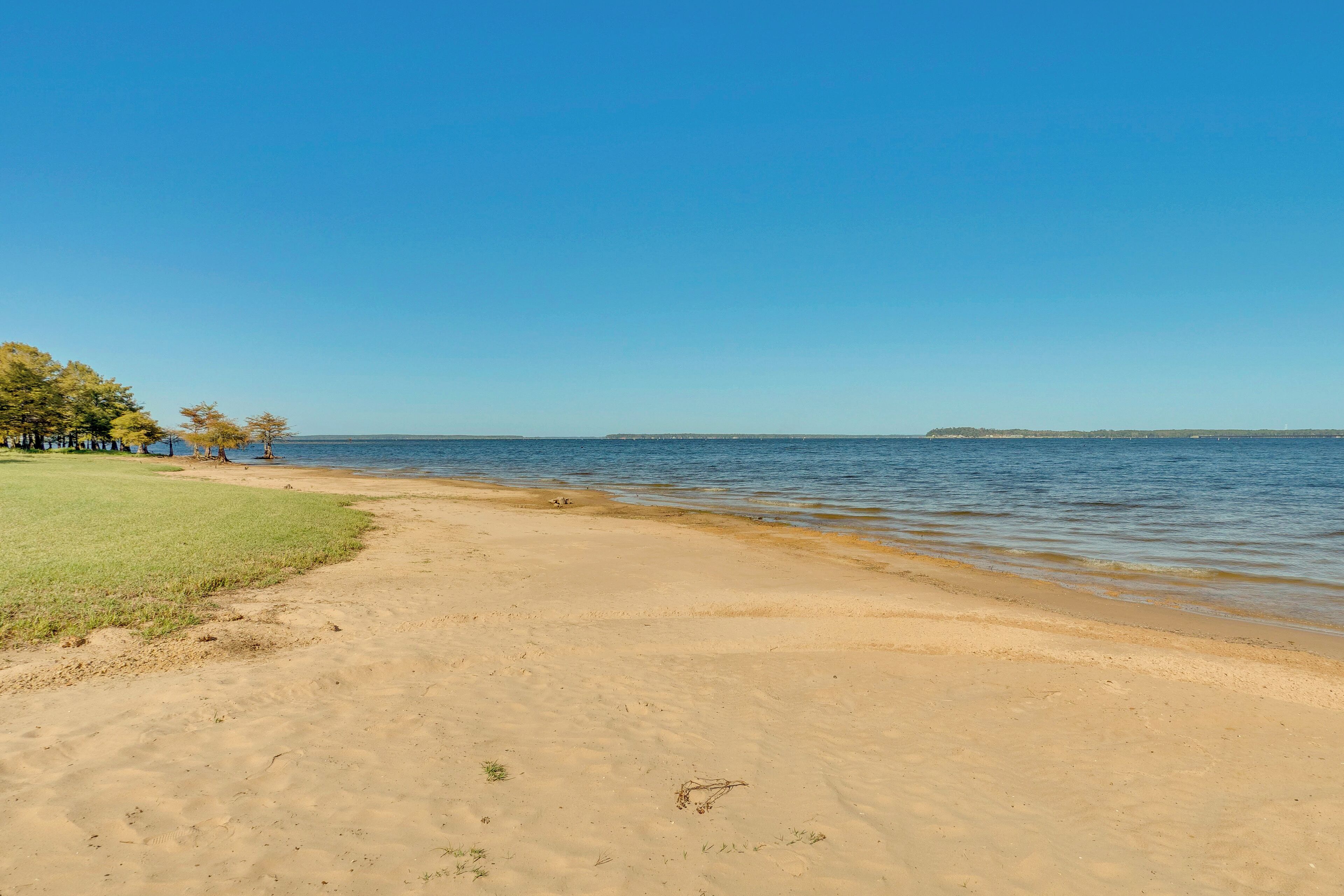 A beach with a clear sky, calm water, and a grassy area with trees.