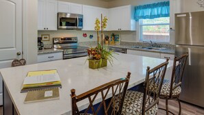A modern kitchen with white cabinets, stainless steel appliances, a central island with chairs, and a window with blue valance. - Game Room + Walk to Beach: Gun Barrel City Gem! (Mabank)