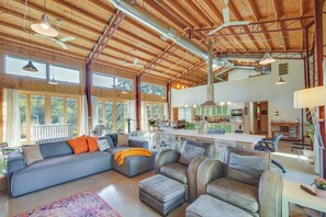A spacious living area with a large sofa, armchairs, and a dining table. There are multiple windows providing natural light and a view of the outdoors.