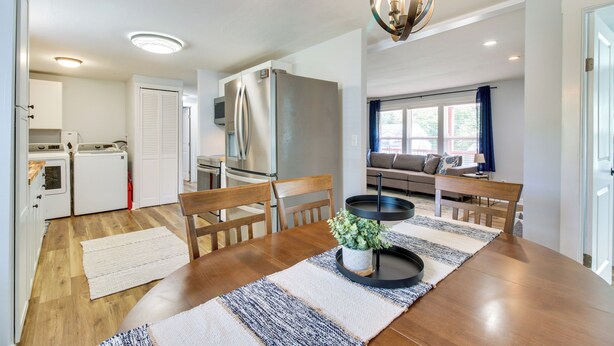 Una cocina moderna con electrodomésticos blancos, un comedor con mesa y sillas de madera, y una sala de estar con sofá y ventana.