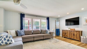Una sala de estar con un sofá modular, un televisor de pantalla plana montado en la pared, un mueble de madera y un ventanal con cortinas.