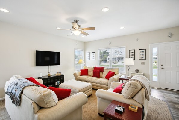 A living room with beige sofas, red cushions, a ceiling fan, a flat-screen TV, and a door leading outside.