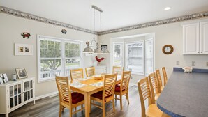 A dining area with a wooden table, chairs, and a large island. There are windows with blinds, a ceiling light, and wall decorations.