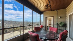 A covered patio with a round table and four chairs, large windows offering a view of the landscape, and a door leading inside.