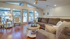 Ein geräumiges Wohnzimmer mit Holzböden, Deckenventilator, großen Fenstern, einem braunen Sofa und einem Couchtisch mit Dekoration.