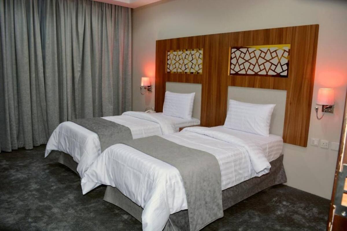 A hotel room with two beds, grey curtains, and wall-mounted lamps.