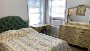 A bedroom with a green tufted headboard, a floral-patterned bedspread, a wooden dresser, and a desk with a chair. - South Lake Charles Guest Cottage - Country Setting, Rustic Ambience (Lake Charles)