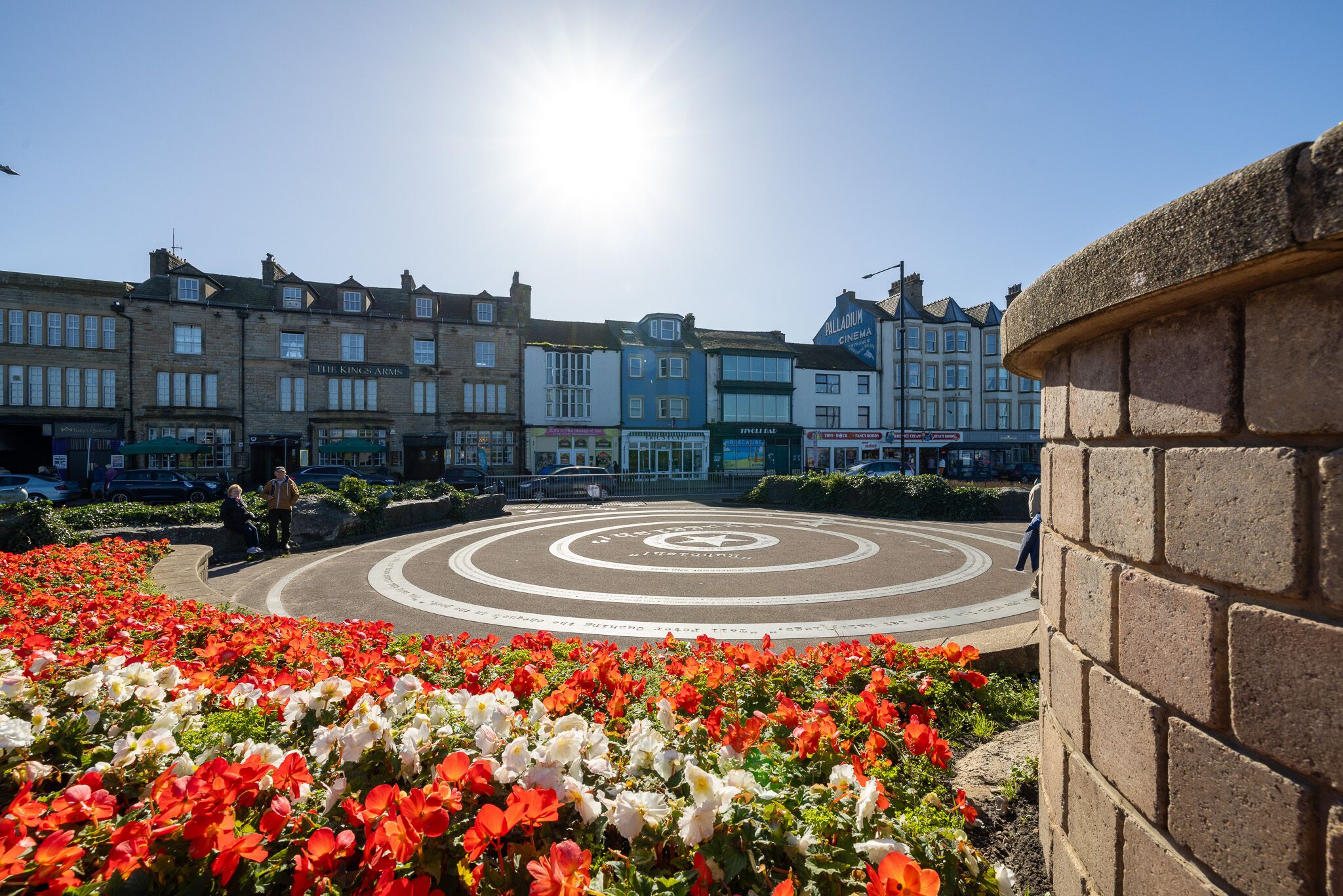 Una plaza pública con un diseño circular en el pavimento, rodeada de macizos de flores y edificios históricos.