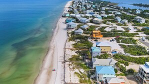 Vue aérienne d’une zone résidentielle côtière, avec des maisons le long de la plage, une eau d’un bleu limpide et un ciel dégagé.